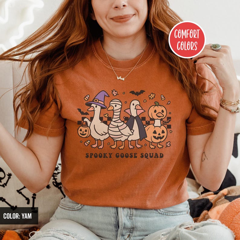 May include: Orange t-shirt with a Halloween design. The graphic shows a "Spooky Goose Squad" with geese in witch, mummy, and vampire costumes, plus a pumpkin. The shirt has the text "COMFORT COLORS" and "COLOR: YAM".