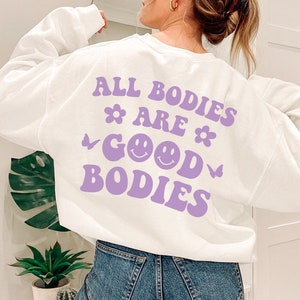 May include: White sweatshirt featuring the phrase "ALL BODIES ARE GOOD BODIES" in purple, with flower, butterfly, and smiley face designs. A relaxed-fit, casual garment.