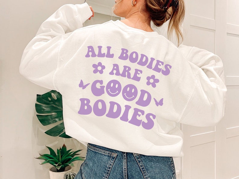 May include: White sweatshirt featuring the phrase "ALL BODIES ARE GOOD BODIES" in purple, with flower, butterfly, and smiley face designs. A relaxed-fit, casual garment.