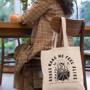 Pu&ograve; includere: Una borsa di tela bianca con testo nero che recita "Books Make Me Feel Alive". La borsa presenta un'illustrazione in bianco e nero di uno scheletro che legge un libro.