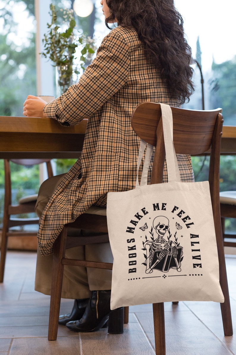 Pu&ograve; includere: Una borsa di tela bianca con testo nero che recita "Books Make Me Feel Alive". La borsa presenta un'illustrazione in bianco e nero di uno scheletro che legge un libro.