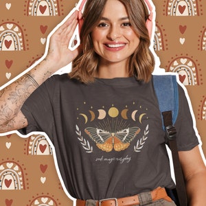 K&ouml;nnte beinhalten: Ein dunkelgraues T-Shirt mit einem Motiv eines Schmetterlings und Mondphasen. Der Text "seek magic everyday" ist unter dem Motiv gedruckt.