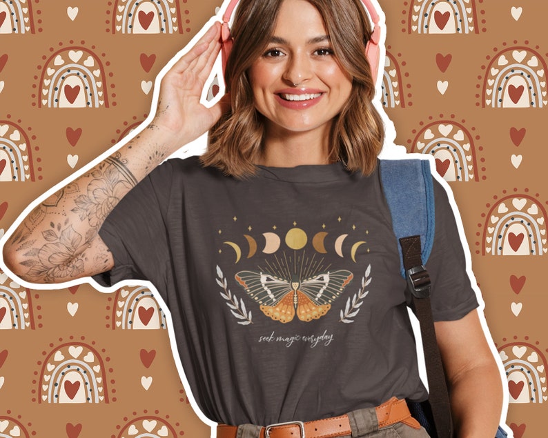 K&ouml;nnte beinhalten: Ein dunkelgraues T-Shirt mit einem Motiv eines Schmetterlings und Mondphasen. Der Text "seek magic everyday" ist unter dem Motiv gedruckt.