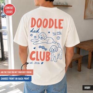 以下が含まれることがあります： 背面に赤い文字でDOODLE DAD CLUBとプリントされたアイボリーのTシャツ。2匹のドゥードル犬、足跡、ボール、おもちゃ、life is better with doodles、est. 2026という文字、小さな星のアイコンが青色で描かれています。