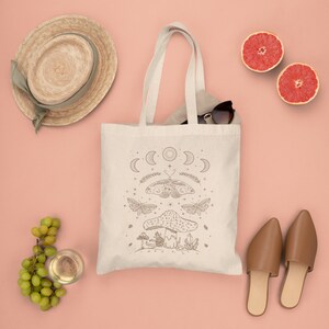 May include: A white canvas tote bag with a brown line drawing of a mushroom, a moth, and a crescent moon design. The bag is on a pink surface with a straw hat, grape clusters, a glass of white wine, and two brown sandals.