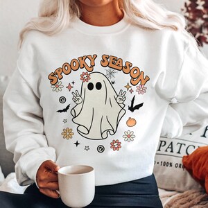 May include: White sweatshirt with a graphic of a ghost with the text "Spooky Season" in orange and yellow letters. The ghost is wearing a peace sign and surrounded by flowers and stars.