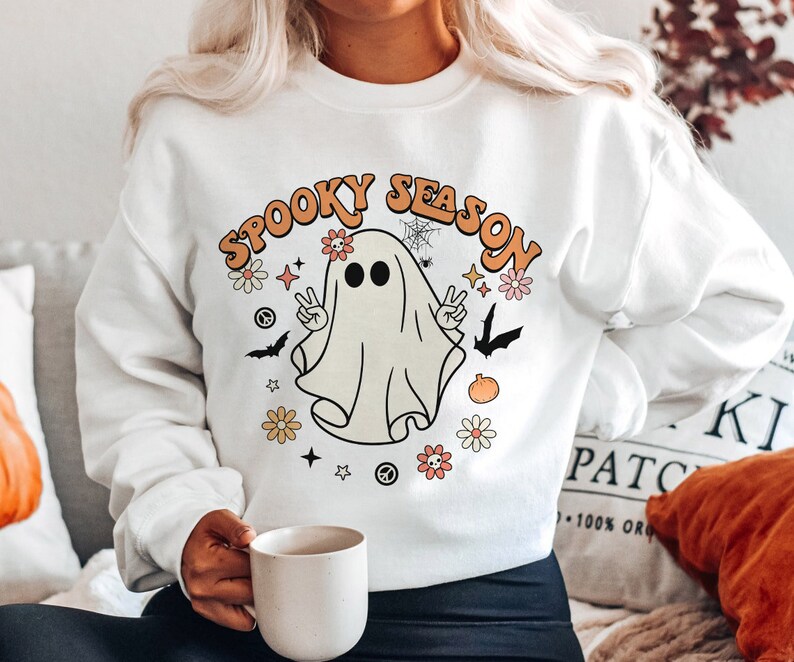 May include: White sweatshirt with a graphic of a ghost with the text "Spooky Season" in orange and yellow letters. The ghost is wearing a peace sign and surrounded by flowers and stars.
