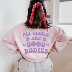 May include: A pale pink sweatshirt featuring the phrase "ALL BODIES ARE GOOD BODIES" in purple, with flower, butterfly, and smiley face graphics. The person is wearing a red bandana in their hair.