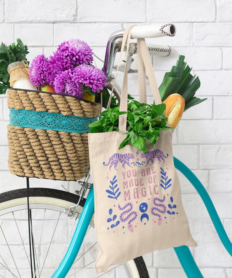 Op de afbeelding: Een beige draagtas met de tekst "YOU ARE MADE OF MAGIC" en een tijger- en slangenontwerp, gevuld met boodschappen, hangt aan een turquoise fiets. Een mand op de fiets bevat paarse bloemen en andere items.