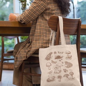 May include: A white tote bag with a brown illustration of mushrooms, frogs, and insects. The bag is hanging on the back of a wooden chair.