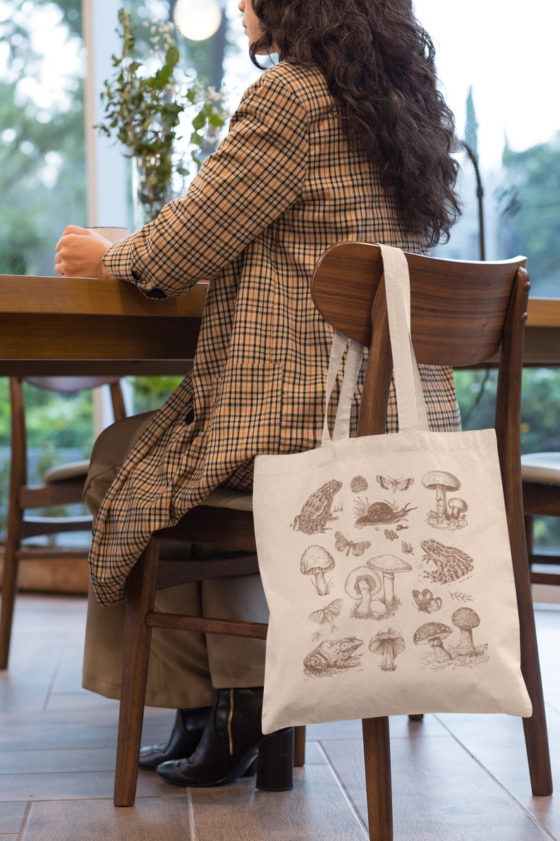 May include: A white tote bag with a brown illustration of mushrooms, frogs, and insects. The bag is hanging on the back of a wooden chair.