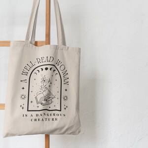 Pode incluir: Bolsa tote bege com texto gr&aacute;fico e ilustra&ccedil;&otilde;es em preto. O texto diz "A WELL-READ WOMAN IS A DANGEROUS CREATURE" acima de um livro aberto com flores e elementos celestes. A bolsa est&aacute; pendurada em um suporte de madeira.