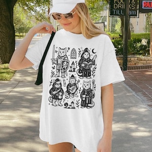 May include: White oversized t-shirt with a black and white graphic print of cats dressed as medieval characters. The graphic includes a knight, a wizard, a bard, and a priest. The shirt is being worn by a person with a white baseball cap and sunglasses.