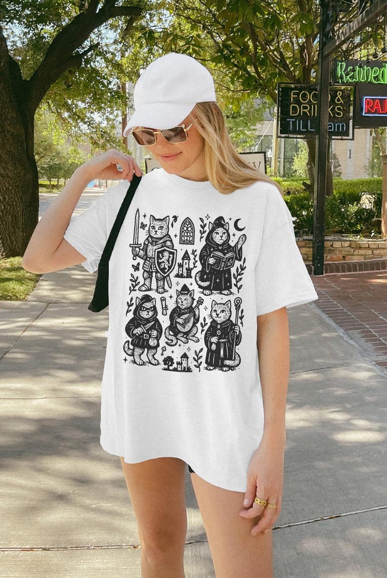 May include: White oversized t-shirt with a black and white graphic print of cats dressed as medieval characters. The graphic includes a knight, a wizard, a bard, and a priest. The shirt is being worn by a person with a white baseball cap and sunglasses.