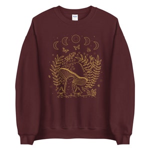 Op de afbeelding: Een maroon sweatshirt met een gouden omtrek van een paddenstoel, maanfasen, vlinders en bladeren. Het ontwerp is gecentreerd op de voorkant van het sweatshirt.