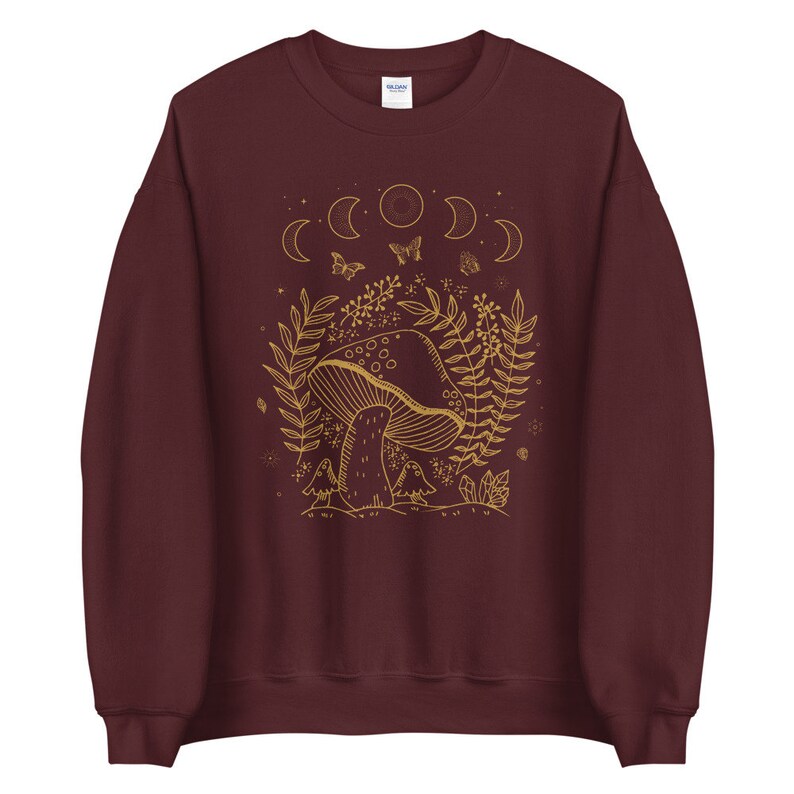 Op de afbeelding: Een maroon sweatshirt met een gouden omtrek van een paddenstoel, maanfasen, vlinders en bladeren. Het ontwerp is gecentreerd op de voorkant van het sweatshirt.