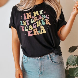May include: Black t-shirt with the text "IN MY 1ST GRADE TEACHER ERA" in colorful, retro-style lettering. The design includes flowers and stars. The shirt is paired with light blue jeans.