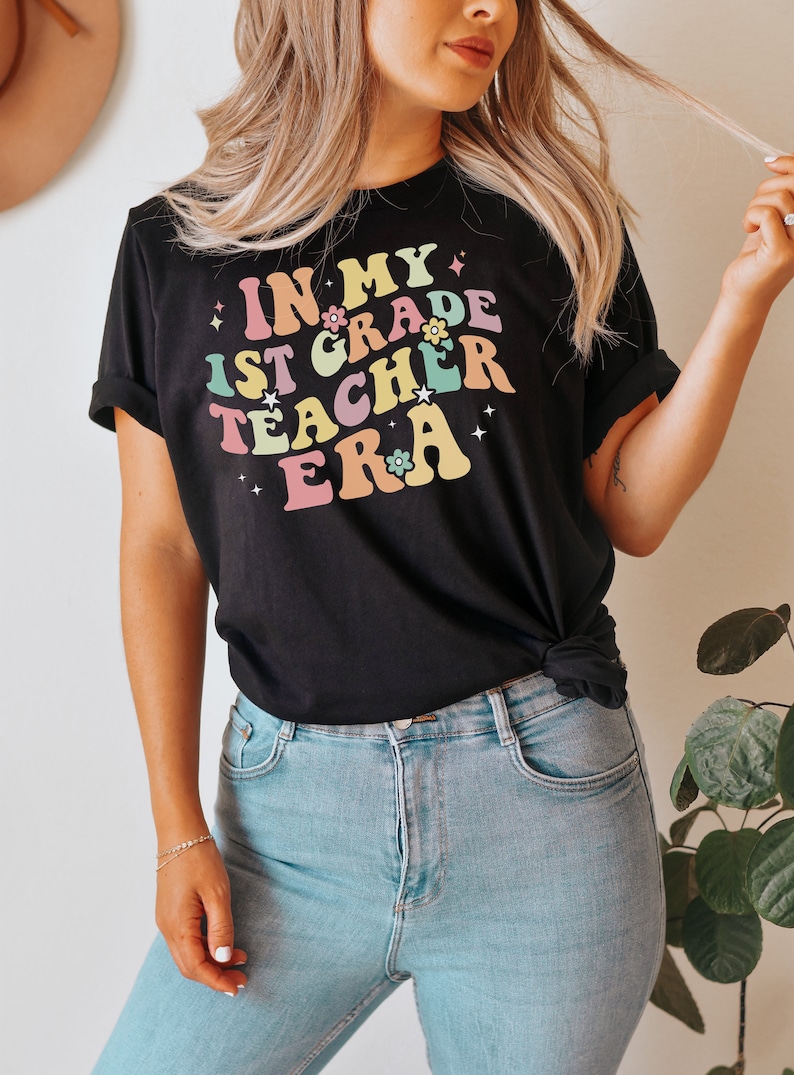 May include: Black t-shirt with the text "IN MY 1ST GRADE TEACHER ERA" in colorful, retro-style lettering. The design includes flowers and stars. The shirt is paired with light blue jeans.