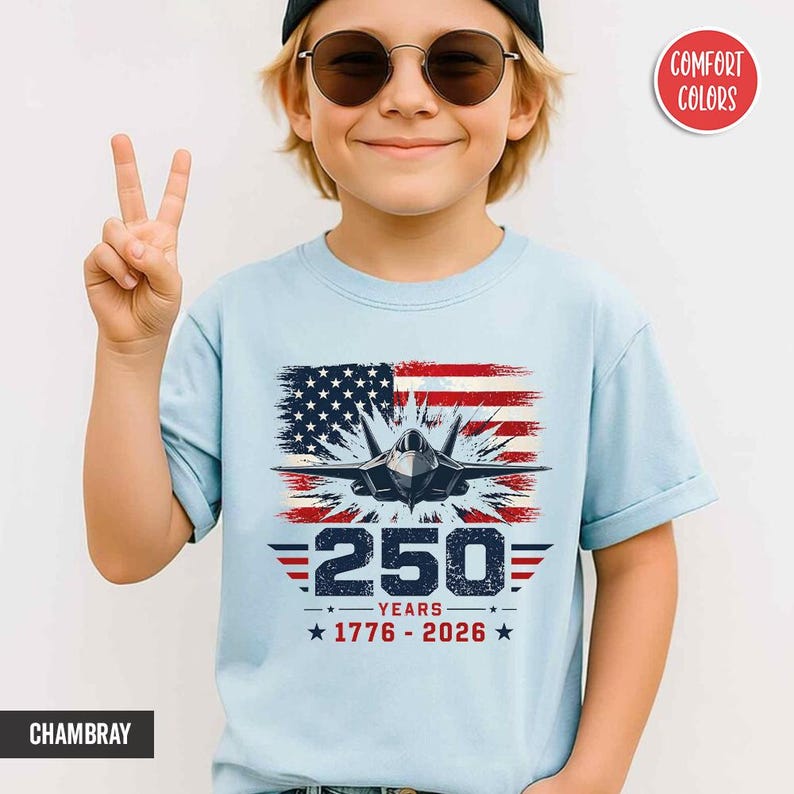 May include: Light blue t-shirt featuring a graphic of an American flag with a jet fighter and the text "250 YEARS 1776-2026". The shirt is made of a soft material and is suitable for everyday wear.