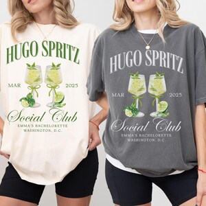 May include: Two white and gray t-shirts with a graphic design of two glasses of Hugo Spritz, a cocktail, with the text "Hugo Spritz" and "Social Club" on the front. The text "MAR 2025" is also on the front of the shirts. The shirts are on two models wearing black shorts.