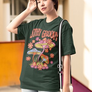 May include: Dark green t-shirt with the words "Stay Groovy" and a colorful mushroom graphic. The design features pink, orange, and blue mushrooms with surrounding flowers. The shirt is paired with light blue jeans and a white purse.