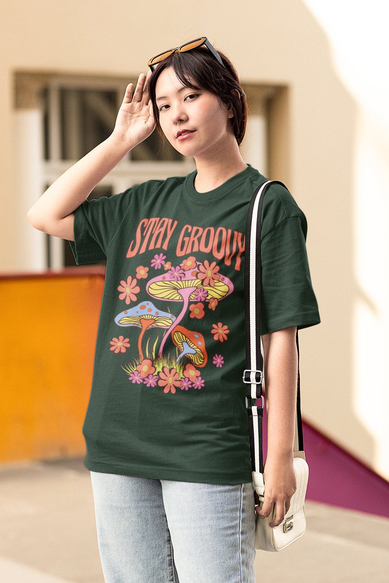 May include: Dark green t-shirt with the words "Stay Groovy" and a colorful mushroom graphic. The design features pink, orange, and blue mushrooms with surrounding flowers. The shirt is paired with light blue jeans and a white purse.