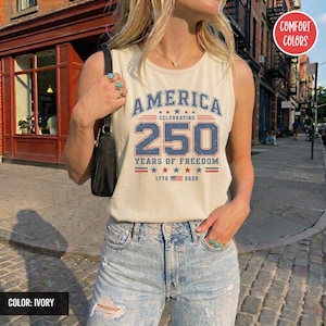 以下が含まれることがあります： アイボリー色のタンクトップ。青と赤で「AMERICA CELEBRATING 250 YEARS OF FREEDOM」の文字がプリントされています。デザインには星と1776年と2026年の文字が含まれています。タンクトップはComfort Colorsブランドです。