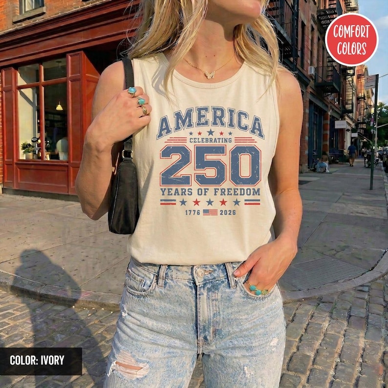 以下が含まれることがあります： アイボリー色のタンクトップ。青と赤で「AMERICA CELEBRATING 250 YEARS OF FREEDOM」の文字がプリントされています。デザインには星と1776年と2026年の文字が含まれています。タンクトップはComfort Colorsブランドです。
