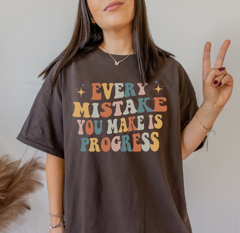May include: A dark brown t-shirt with the text "EVERY MISTAKE YOU MAKE IS PROGRESS" in colorful, retro-style lettering. The design includes stars and a vintage font, creating a motivational and positive message.