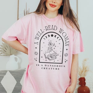 May include: A pink t-shirt with a black graphic design that reads "A well-read woman is a dangerous creature". The graphic features a stack of books, a flower, and a crescent moon.