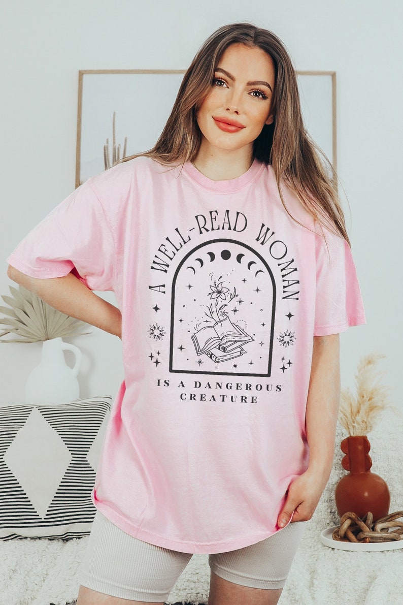 May include: A pink t-shirt with a black graphic design that reads "A well-read woman is a dangerous creature". The graphic features a stack of books, a flower, and a crescent moon.