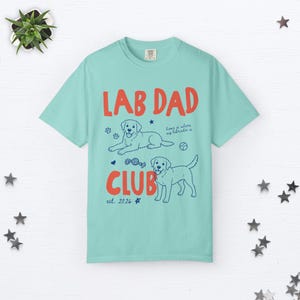 Puede incluir: Una camiseta de manga corta de color verde azulado claro con el texto rojo LAB DAD CLUB y el texto azul est. 2026. El gr&aacute;fico incluye dos ilustraciones de Labrador, huellas, una pelota, un coraz&oacute;n y el texto home is where my labrador is.