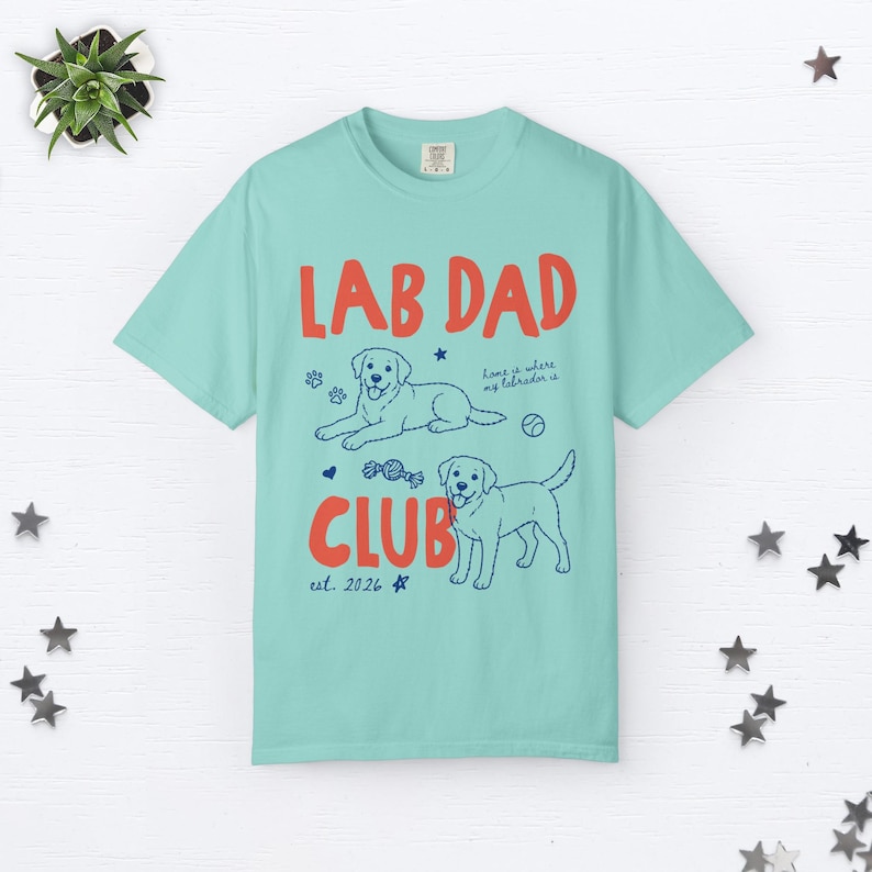 Puede incluir: Una camiseta de manga corta de color verde azulado claro con el texto rojo LAB DAD CLUB y el texto azul est. 2026. El gr&aacute;fico incluye dos ilustraciones de Labrador, huellas, una pelota, un coraz&oacute;n y el texto home is where my labrador is.
