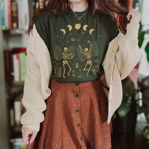 May include: A dark green t-shirt with a graphic of two skeletons dancing with wings. The skeletons are gold and the background is a dark green with gold stars and moons. The shirt is worn with a cream cardigan and a brown corduroy skirt.