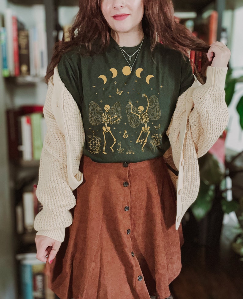 May include: A dark green t-shirt with a graphic of two skeletons dancing with wings. The skeletons are gold and the background is a dark green with gold stars and moons. The shirt is worn with a cream cardigan and a brown corduroy skirt.