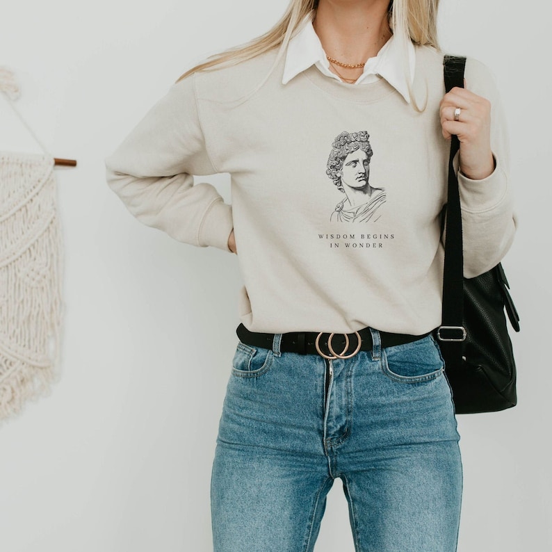 May include: A beige sweatshirt with a graphic of a man's face and the text "Wisdom begins in wonder".