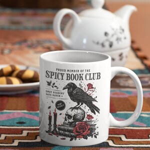 Puede incluir: Taza de cer&aacute;mica blanca con una ilustraci&oacute;n en blanco y negro de un cuervo posado sobre un cr&aacute;neo con una rosa roja. La taza dice "Proud Member of the Spicy Book Club" y "Smut Readers' Safe Haven + Meet Weekly".
