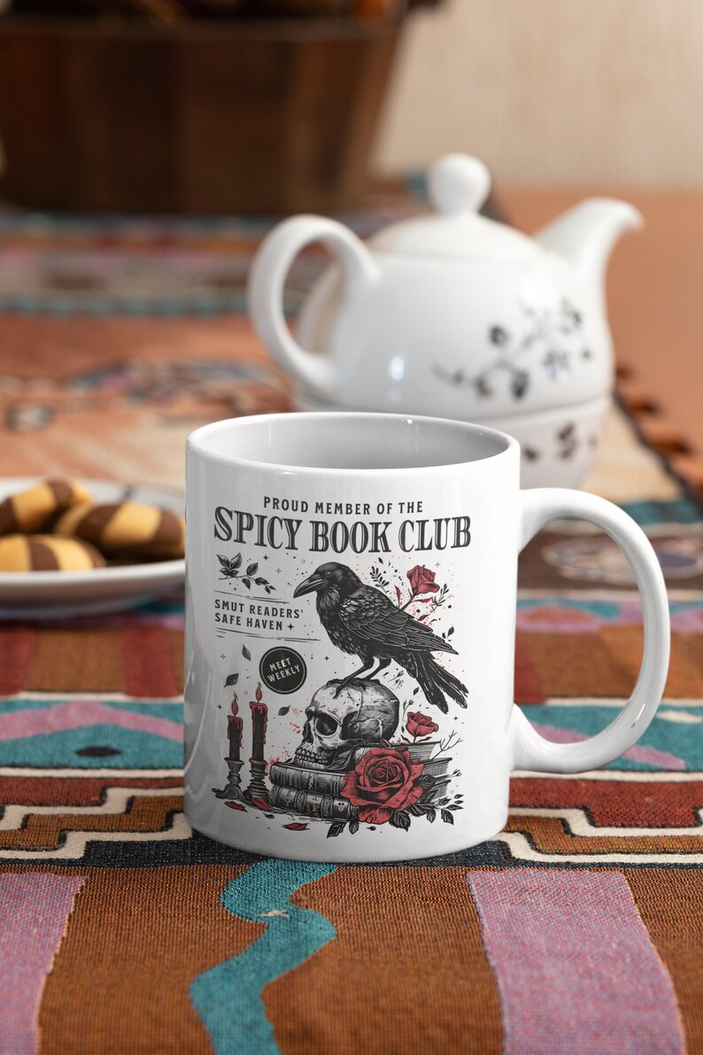 Puede incluir: Taza de cer&aacute;mica blanca con una ilustraci&oacute;n en blanco y negro de un cuervo posado sobre un cr&aacute;neo con una rosa roja. La taza dice "Proud Member of the Spicy Book Club" y "Smut Readers' Safe Haven + Meet Weekly".