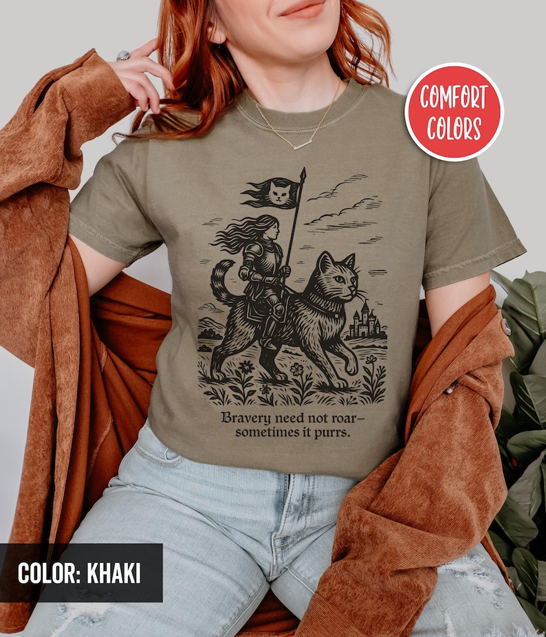 Peut inclure: Un t-shirt kaki avec une illustration en noir et blanc d'une femme chevauchant un chat. Le chat tient un drapeau avec un visage de chat dessus. Le texte sur le t-shirt est "Bravery need not roar - sometimes it purrs."