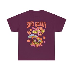 May include: A maroon t-shirt featuring a colorful graphic with the text "STAY GROOVY". The graphic includes stylized mushrooms, flowers, and grass in shades of yellow, pink, orange, and blue.