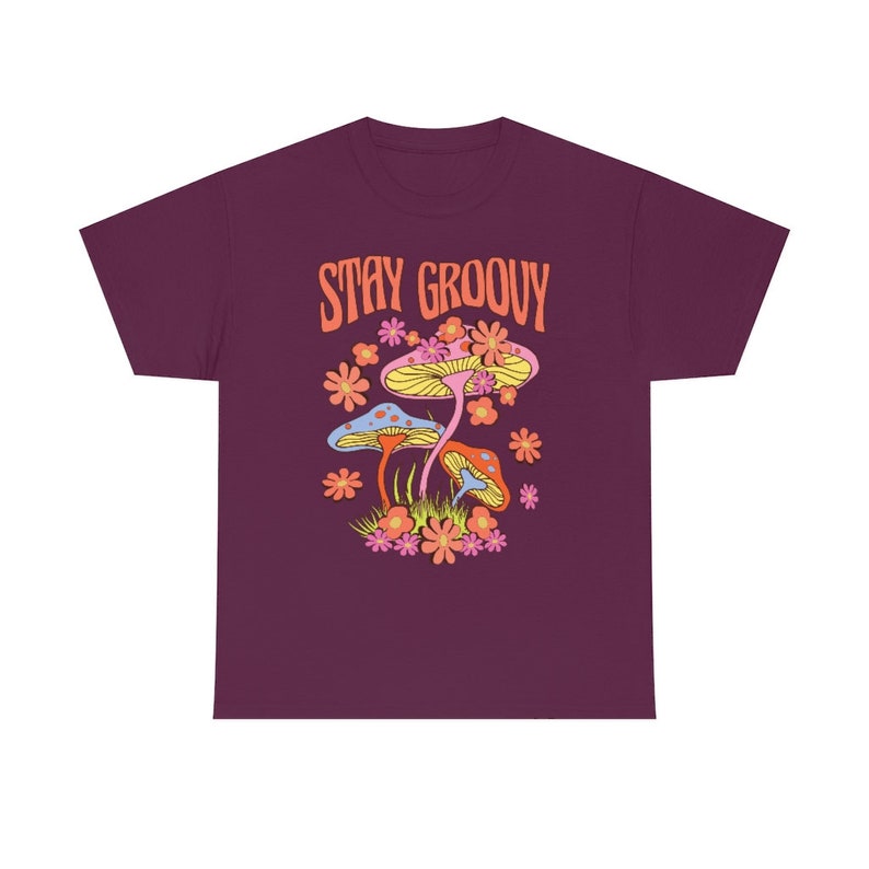 May include: A maroon t-shirt featuring a colorful graphic with the text "STAY GROOVY". The graphic includes stylized mushrooms, flowers, and grass in shades of yellow, pink, orange, and blue.