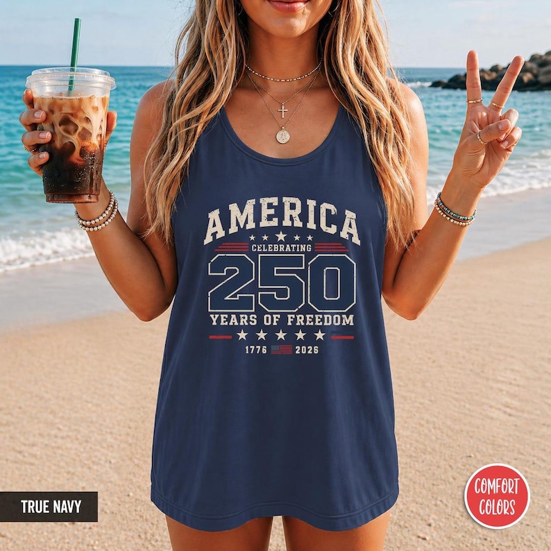 以下が含まれることがあります： 白と赤で「AMERICA CELEBRATING 250 YEARS OF FREEDOM 1776-2026」の文字が入ったネイビーブルーのタンクトップ。タンクトップはComfort Colorsブランドです。画像はビーチで撮影されています。