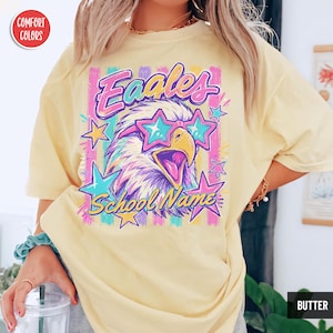 May include: A butter yellow short-sleeve t-shirt featuring a graphic of an eagle wearing star-shaped sunglasses. The design includes the text "Eaales" and "School Name" surrounded by colorful vertical stripes and various star shapes in pink and blue.