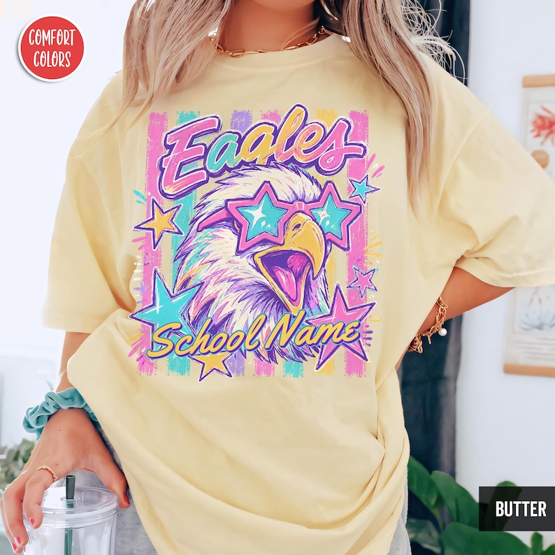 May include: A butter yellow short-sleeve t-shirt featuring a graphic of an eagle wearing star-shaped sunglasses. The design includes the text "Eaales" and "School Name" surrounded by colorful vertical stripes and various star shapes in pink and blue.