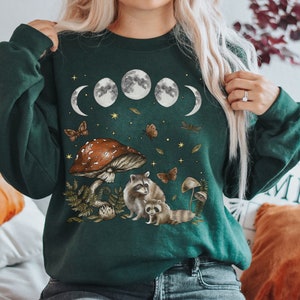 May include: A dark green sweatshirt with a moon phase design, mushrooms, and raccoons. The design is in shades of brown, white, and gray. The sweatshirt has a crew neck and long sleeves.