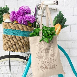 May include: A beige tote bag with a moon and butterfly design. The bag is hanging on a bicycle with a wicker basket filled with flowers and groceries.