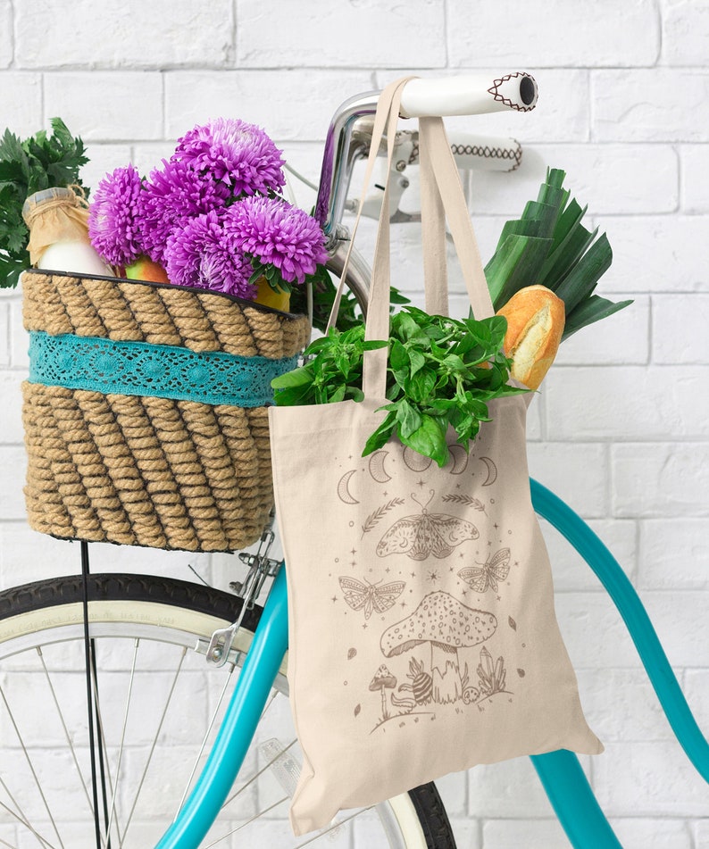 May include: A beige tote bag with a moon and butterfly design. The bag is hanging on a bicycle with a wicker basket filled with flowers and groceries.