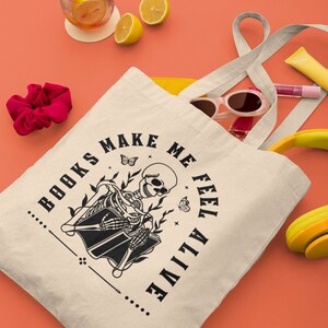 Pu&ograve; includere: Una borsa di tela bianca con testo nero che recita "...BOOKS MAKE ME FEEL ALIVE..." e un'illustrazione in bianco e nero di uno scheletro che legge un libro. La borsa &egrave; appoggiata su una superficie color corallo con altri oggetti come occhiali da sole, un elastico per capelli e una bevanda gialla con fette di limone.
