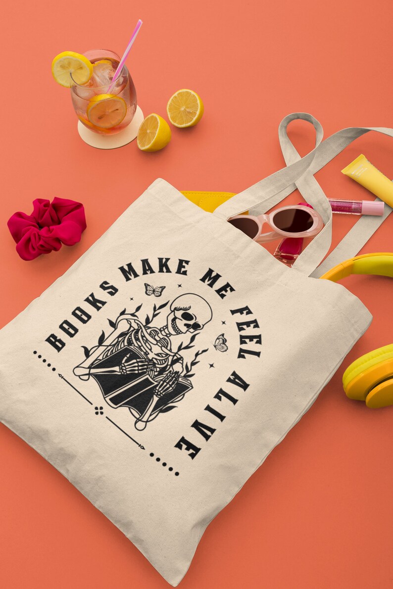 Pu&ograve; includere: Una borsa di tela bianca con testo nero che recita "...BOOKS MAKE ME FEEL ALIVE..." e un'illustrazione in bianco e nero di uno scheletro che legge un libro. La borsa &egrave; appoggiata su una superficie color corallo con altri oggetti come occhiali da sole, un elastico per capelli e una bevanda gialla con fette di limone.