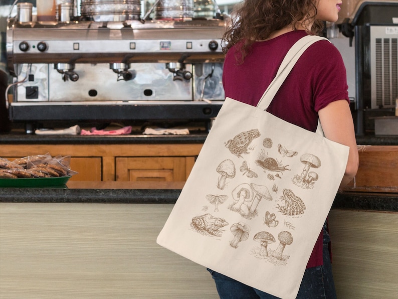 May include: A beige tote bag with a brown illustration of mushrooms, frogs, butterflies, and other forest creatures. The bag has two handles and is being held by a person.